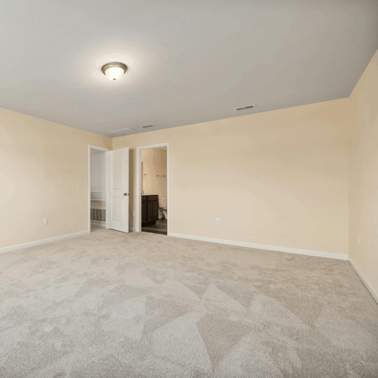 A spacious empty room featuring beige walls and plush carpet, with an open door leading to a bathroom.